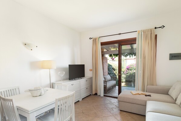 Una habitación con una mesa blanca, un televisor sobre un aparador, un sofá y un ventanal corredero que da a un espacio exterior.