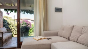 A cozy living room with a beige sofa, a wicker chair, and a glass door leading to a balcony with a view of greenery and a building. - Casa di Barbara al Mare by Wonderful Italy (Olbia)