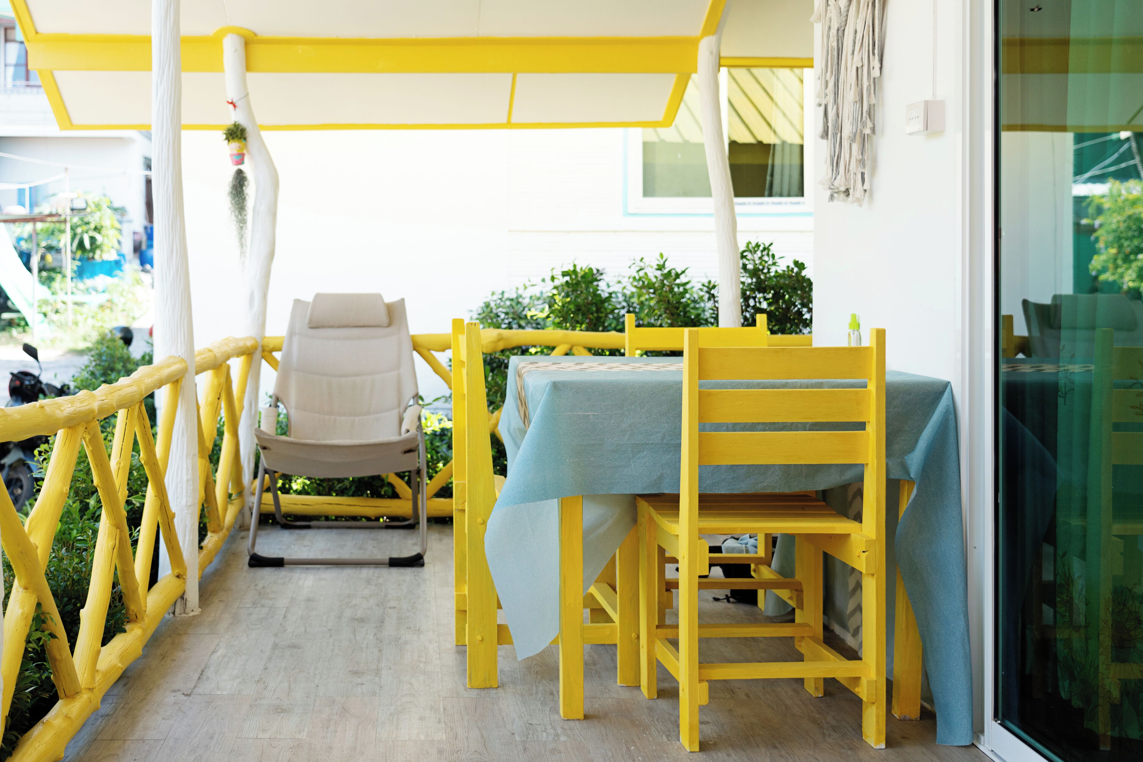A yellow outdoor seating area with a table and chairs, a wooden railing, and a view of greenery.