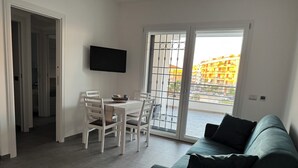 A living room with a dining area, a TV mounted on the wall, and a sliding glass door leading to a balcony with a view of buildings.