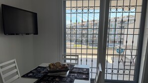 A dining area with a table set for two, a TV mounted on the wall, and a view through a barred window to the outside.