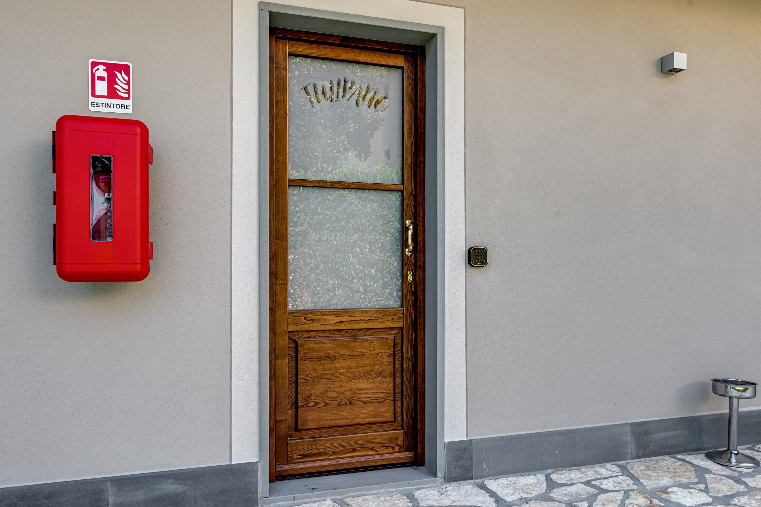 Una puerta de madera con un panel de vidrio esmerilado, un letrero de 'Welcome', una caja roja contra incendios y una lámpara exterior de metal.