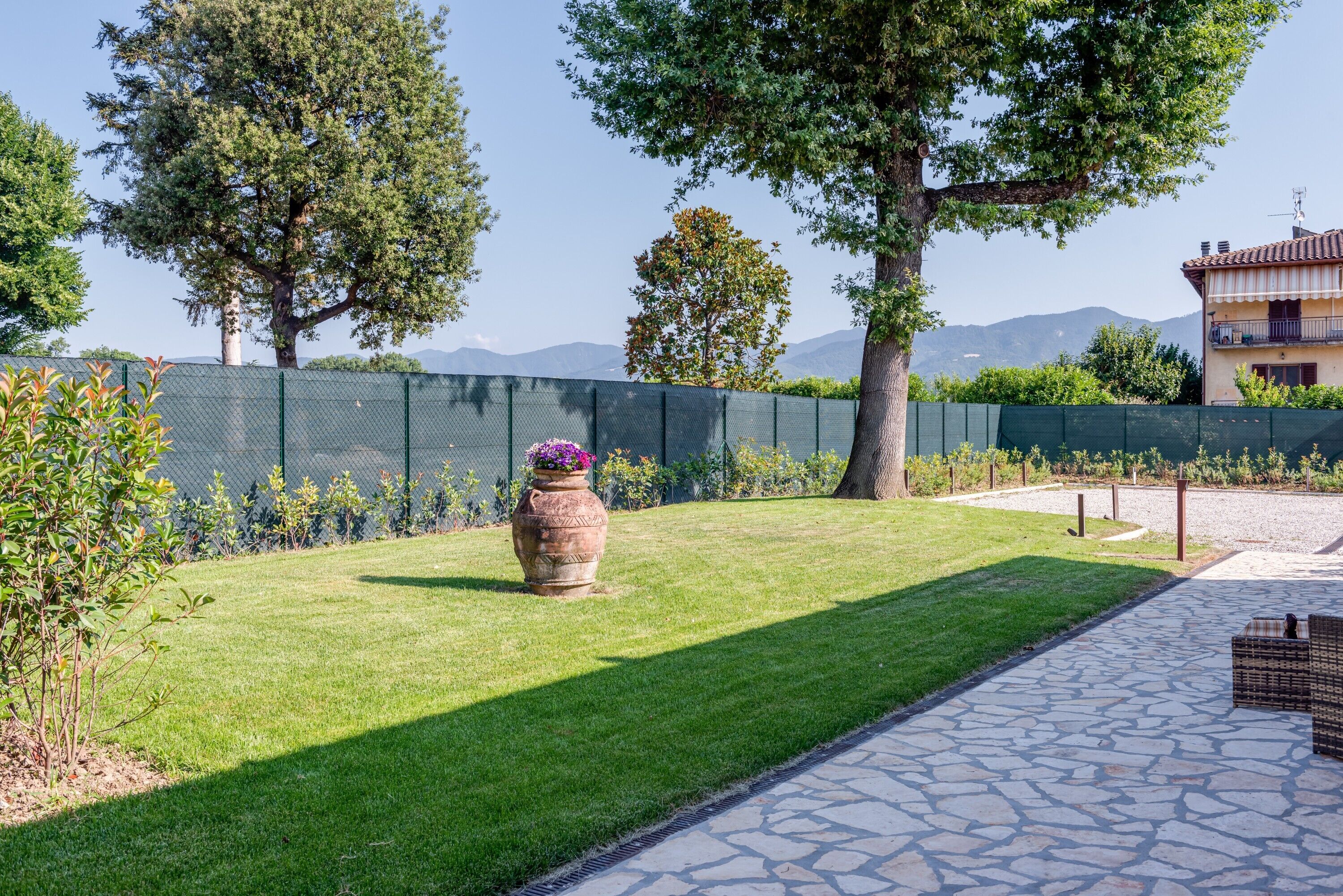 Un césped bien cuidado con un sendero de piedra, una gran urna decorativa y una cerca verde.