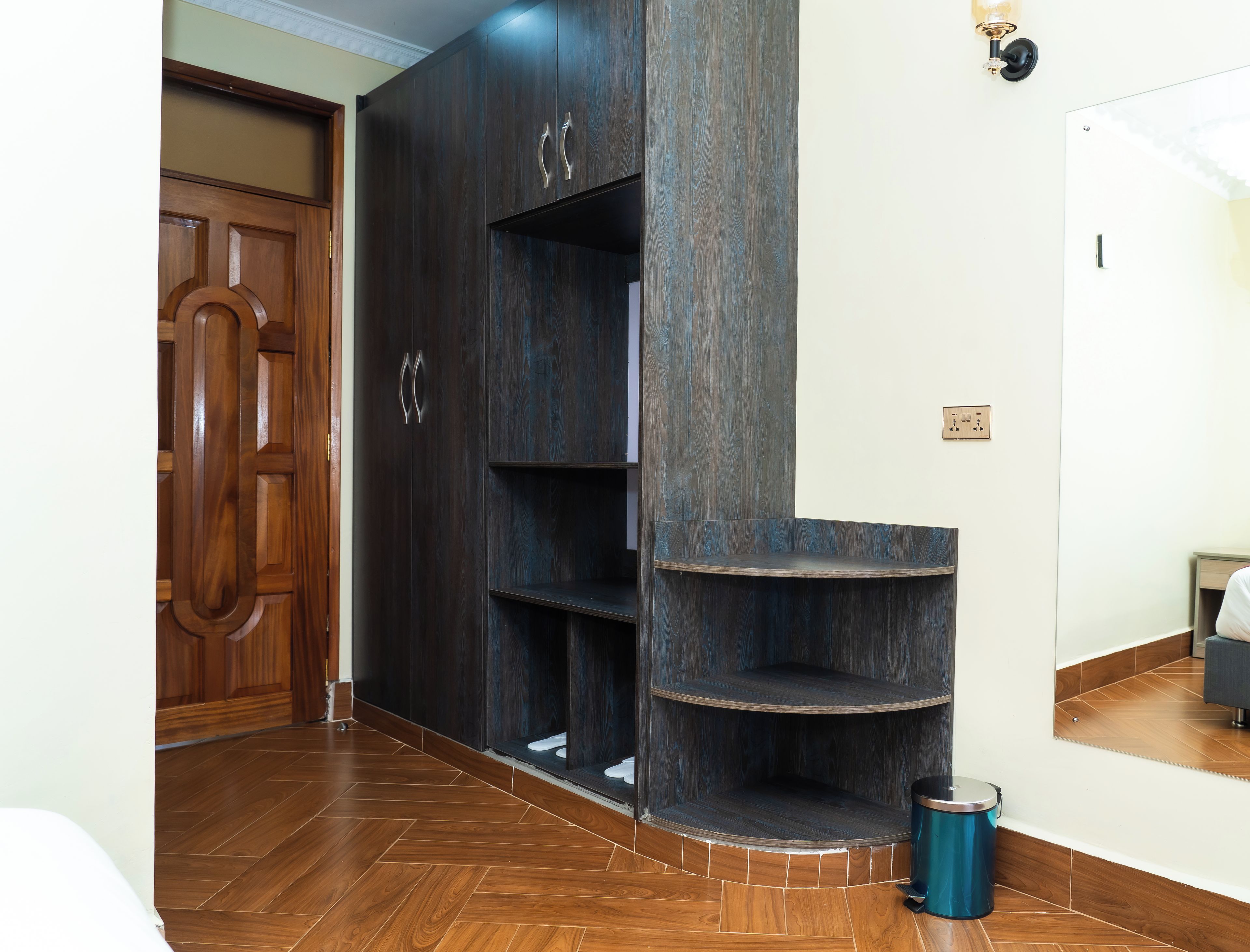 Una habitación con un armario de madera, una estantería empotrada, un cesto de basura y una cama con una mesita de noche.