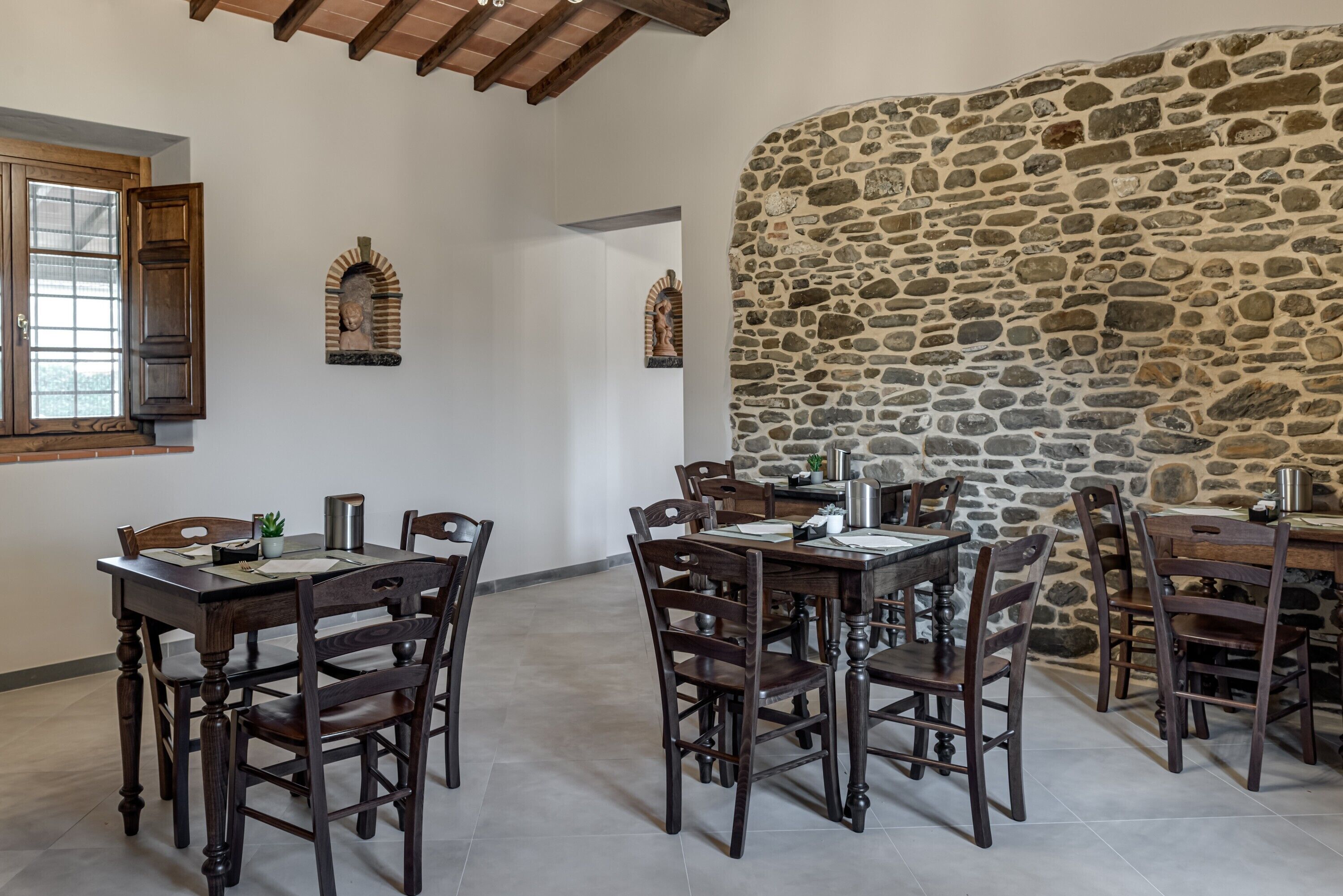 A dining area with wooden tables and chairs, stone walls, and arched niches with sculptures.
