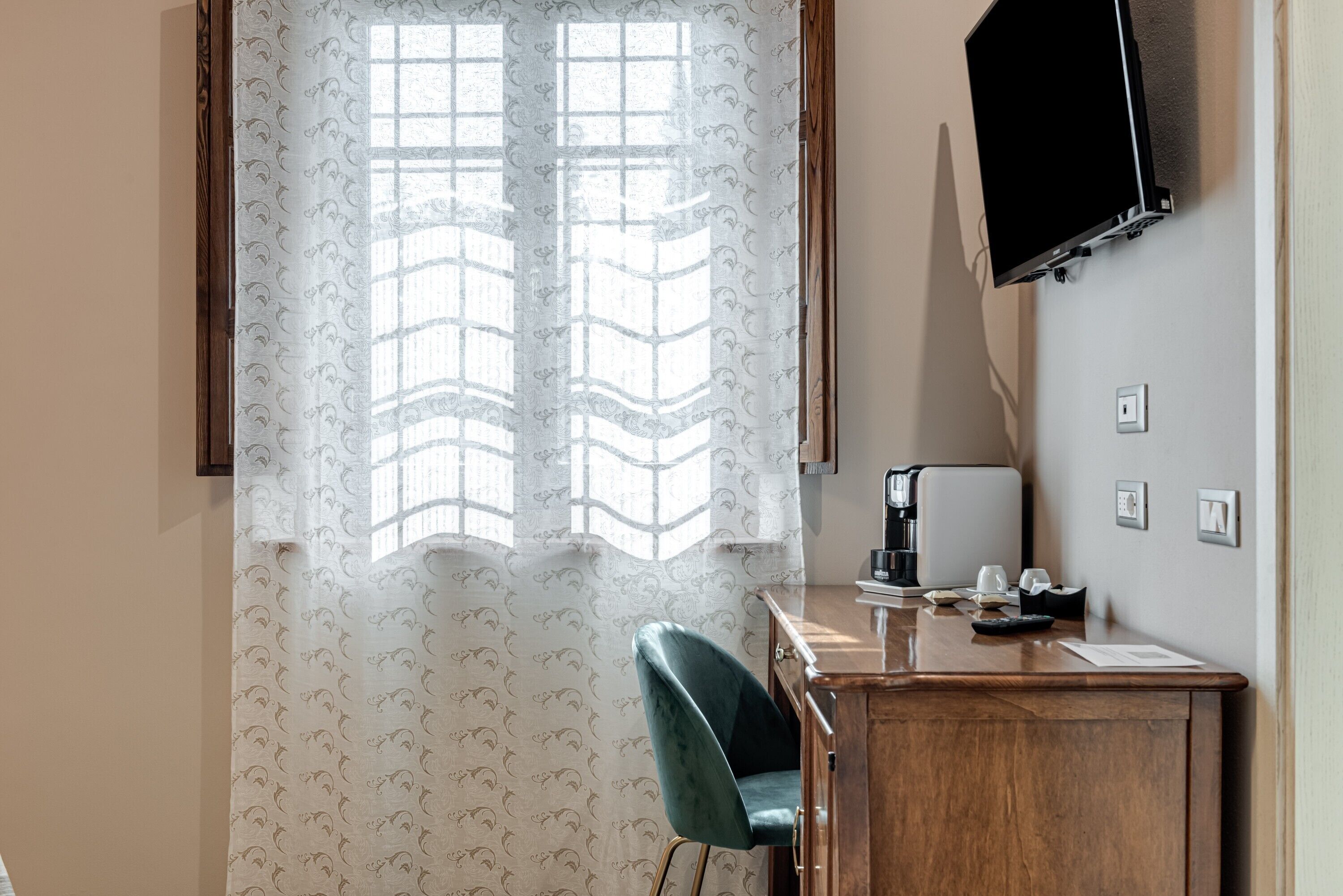 Une pièce comprenant un bureau, une chaise, un téléviseur fixé au mur et une fenêtre ornée de rideaux à motifs.