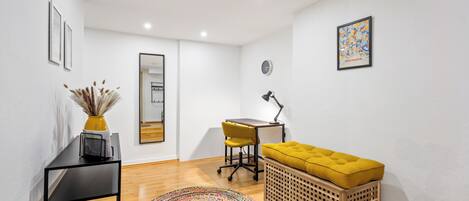 A modern, well-lit room with a wooden floor, a black desk, a yellow chair, a yellow ottoman, a colorful rug, and a wooden basket.