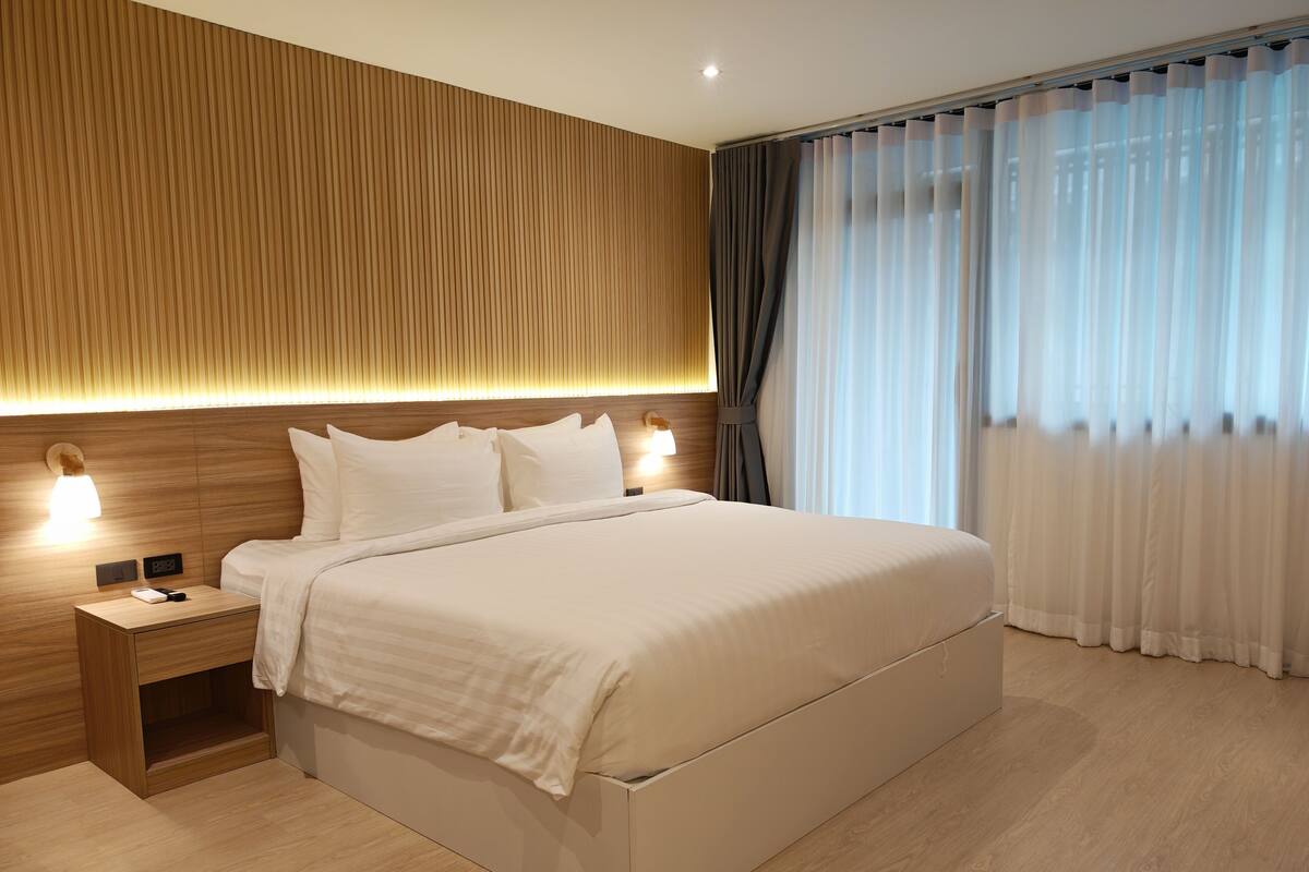 A modern hotel room with a large bed, wooden headboard, bedside lamps, and a view of the outdoors through sheer curtains.