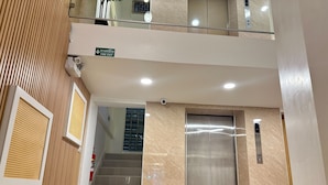 A modern elevator with a metallic finish, a fire exit sign above the stairs, and a textured wall with a vertical pattern. - Winston Hotel (Bangkok)