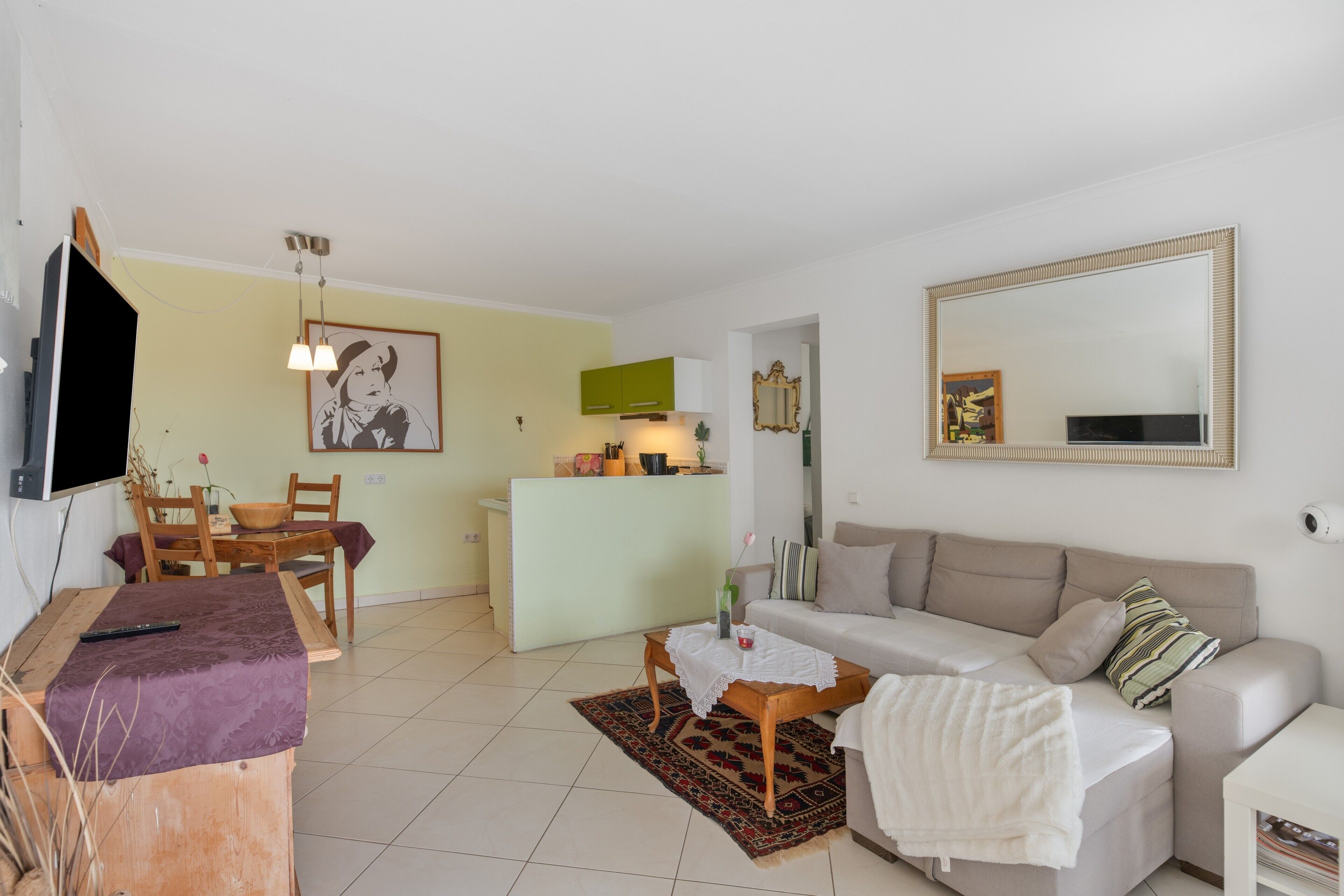 A living room with a sofa, a coffee table, a TV, a dining area with chairs, a kitchen area with a counter, and a large mirror on the wall.