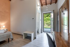 A modern hotel room with a bed, bedside table, bench, and a view of the garden through an open door.