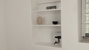 A minimalist room with a built-in white shelving unit against the wall, a small bedside table, and a window with blinds.