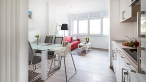 A modern kitchen with white cabinets and a stainless steel refrigerator. There is a dining area with a glass table and chairs. A red sofa is visible in the background. - // KING APARTMENTS - APARTMENT 2 // (Norderney)