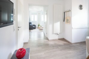 A hallway with a red apple on a mat, a coat rack with bags, and a door leading to a living area with a sofa and a plant. - // KÖNIG APARTMENTS - APARTMENT 3 // (Norderney)