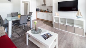 A modern living room with a glass dining table, a red sofa, a white coffee table with a magazine, and a TV mounted on the wall. - // KING APARTMENTS - APARTMENT 5 // (Norderney)