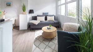 Moderni olohuone, jossa on harmaa sohva, pyöreä sohvapöytä ja suuret ikkunat, joista on näkymä ulos.