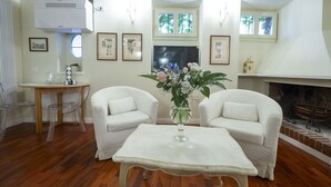 Un salon avec deux fauteuils blancs, une petite table et un vase rempli de fleurs. On y trouve une cheminée, des tableaux encadrés au mur et une fenêtre donnant sur des arbres.