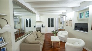 Un vaste salon avec un canapé beige, un fauteuil blanc et une table basse. On aperçoit, au fond, une cuisine avec des meubles blancs et une cuisinière.