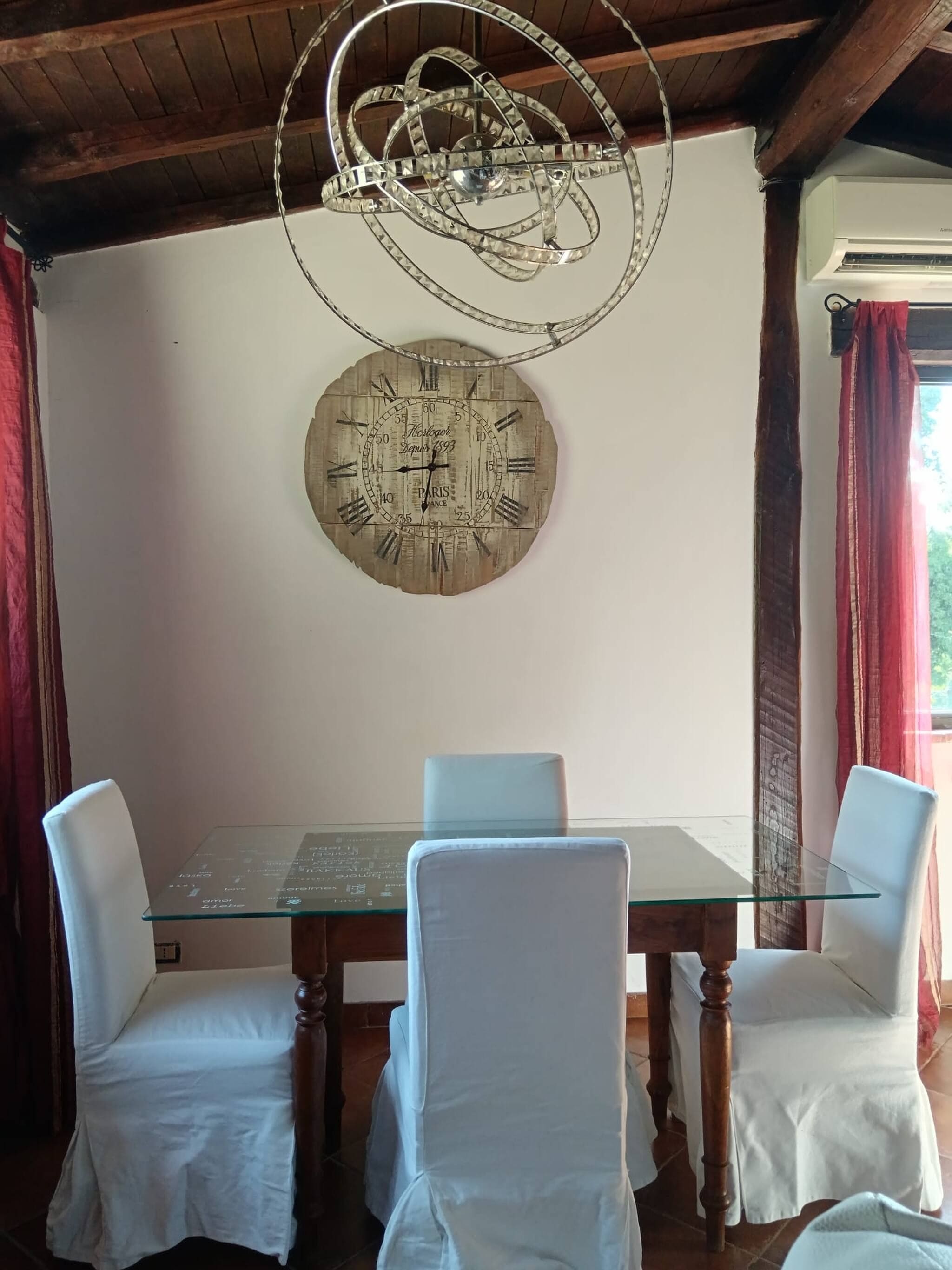 Een eetgedeelte met een glazen tafel, witte stoelen, een houten plafond en een decoratieve klok aan de muur.