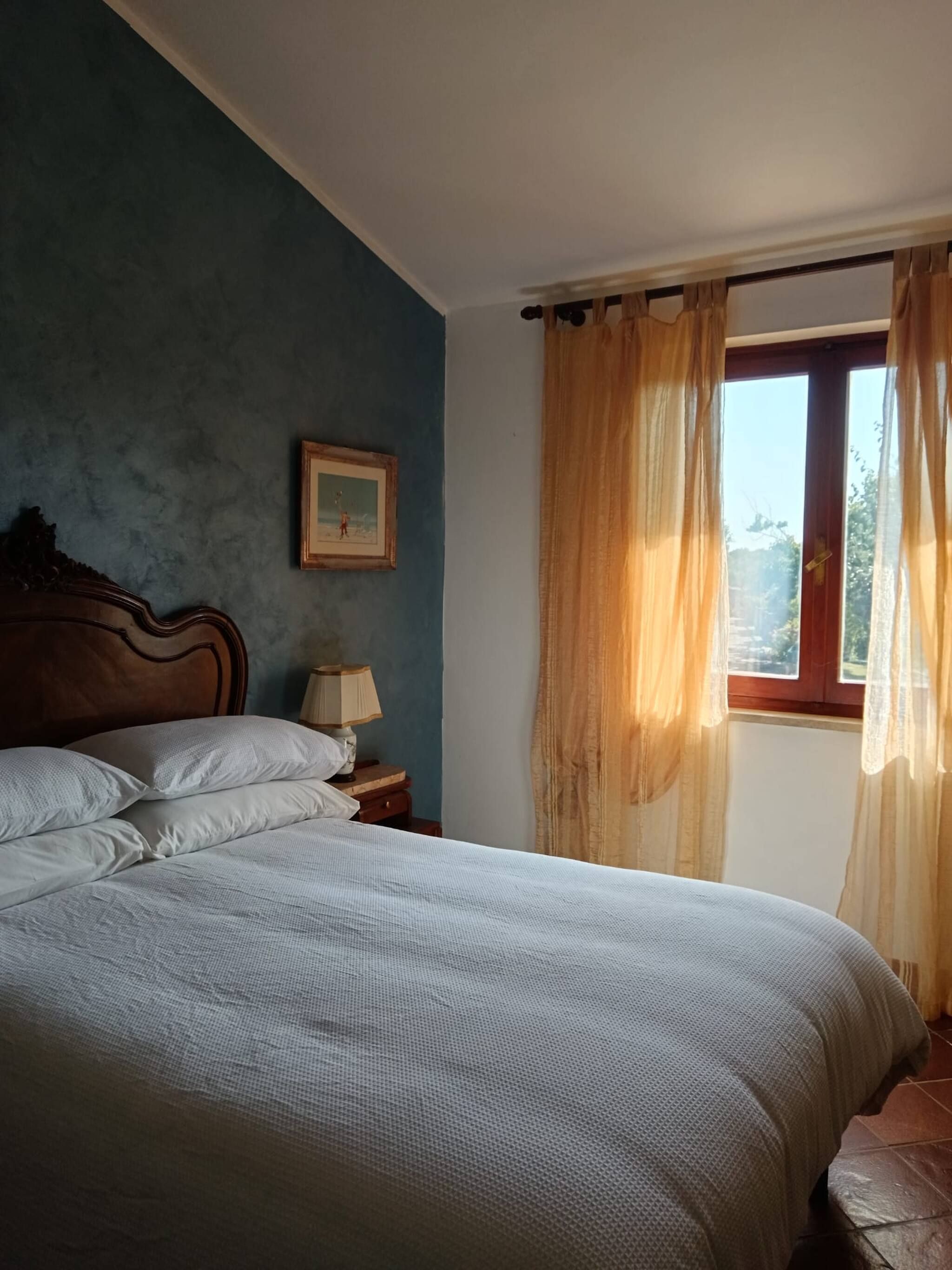 A bedroom with a bed, a nightstand, a lamp, a framed picture on the wall, and a window with curtains.