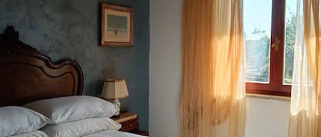 Een slaapkamer met een bed, nachtkastje, lamp, een ingelijste foto aan de muur en een raam met gordijnen.