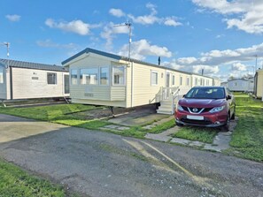 A mobile home with a car parked in front, a grassy yard, and a clear sky.