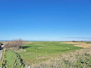 Un parcours de golf sous un ciel dégagé, avec un sentier sur la gauche et des éoliennes visibles au loin.