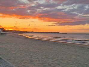 A beach with a sunset, waves, and a distant shoreline with buildings.