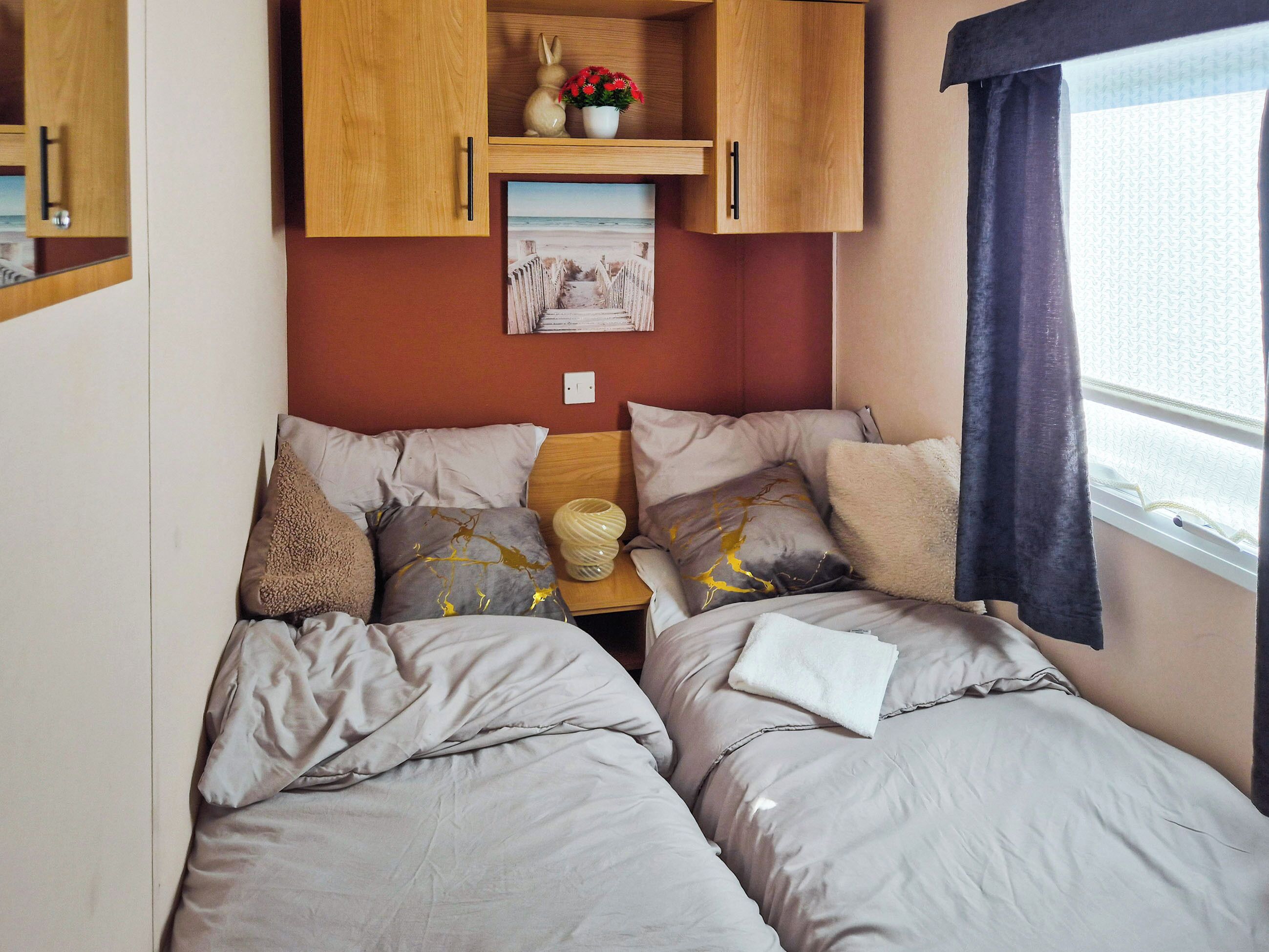 Een slaapkamer met twee bedden, houten kasten erboven, een klein tafeltje met een vaas, en een raam met gordijnen.