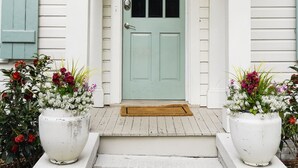 Una casa blanca con una puerta verde, un cartel sobre la puerta y dos macetas grandes en los escalones.