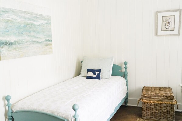 A single bed with a blue headboard, a white wall with vertical paneling, a small wicker basket, and a framed picture on the wall.