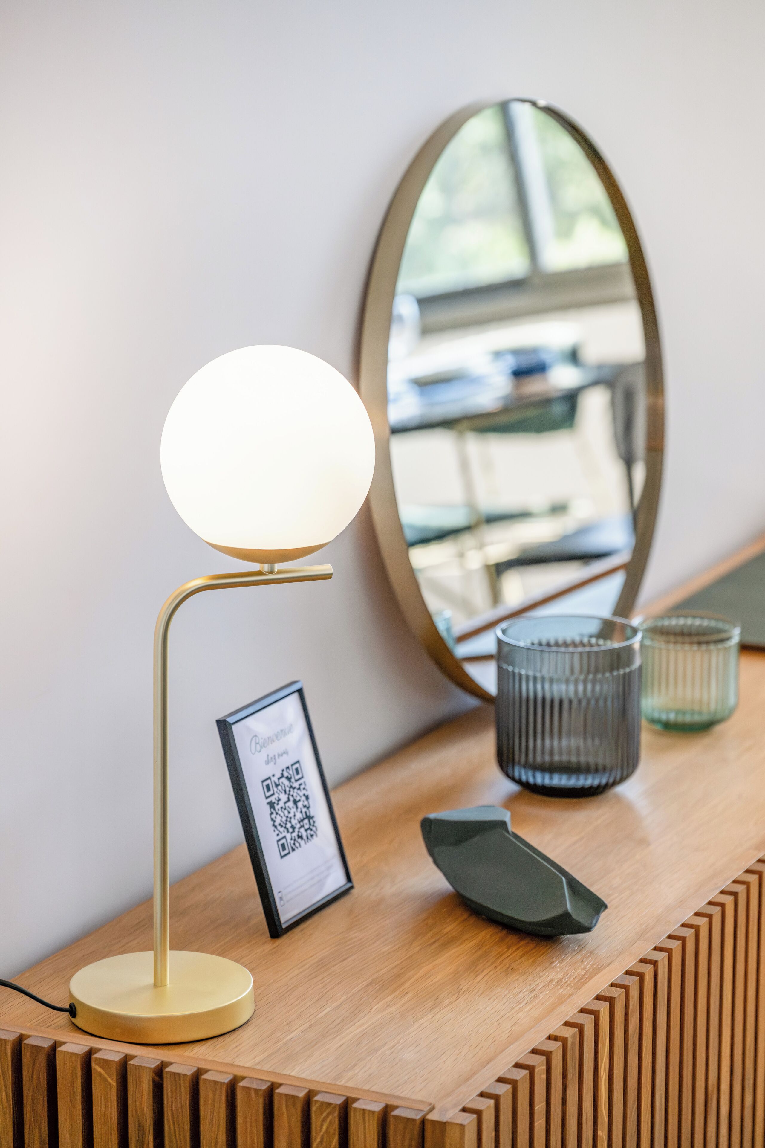 A desk with a round table lamp, a framed QR code, a mirror, and two glass vases.
