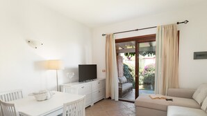 Une pièce comprenant une table à manger blanche, un canapé, un téléviseur posé sur une commode et une porte-fenêtre coulissante donnant sur un espace extérieur.