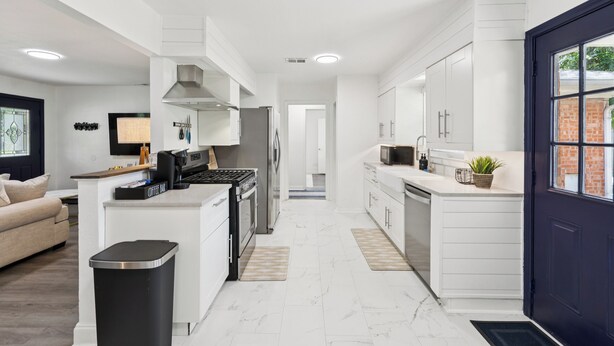 A modern kitchen with white cabinetry, a black trash bin, and a blue door. - Boho Bungalow near DFW Airport (Farmers Branch)