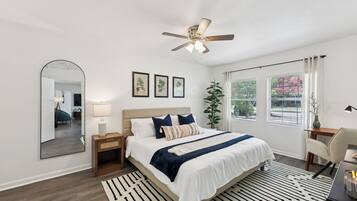 A modern bedroom with a large bed, a ceiling fan, a mirror, and a view of the outdoors through large windows.