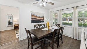 A dining area with a wooden table, chairs, a ceiling fan, and a view of the outdoors through large windows. - Boho Bungalow near DFW Airport (Farmers Branch)
