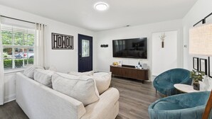 A modern living room with a beige sofa, a blue armchair, a TV, and a wooden floor. - Boho Bungalow near DFW Airport (Farmers Branch)