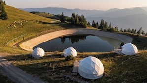 Un espacio al aire libre circular con un estanque, rodeado por una terraza de madera y tres estructuras en forma de cúpula.