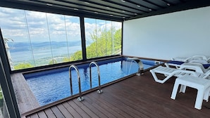 An indoor swimming pool with clear views of a lake and mountains, surrounded by wooden decking and white lounge chairs. - adesa viev villas (Sapanca)