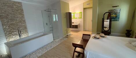 A modern bathroom with a bathtub, shower, and glass-enclosed shower area. There is a wooden chair and a bed with a white bedspread.