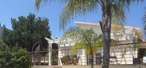 A building with a white facade, a covered patio area with outdoor furniture, and a palm tree in the foreground. - Castillitos en el Aire  (Denia)