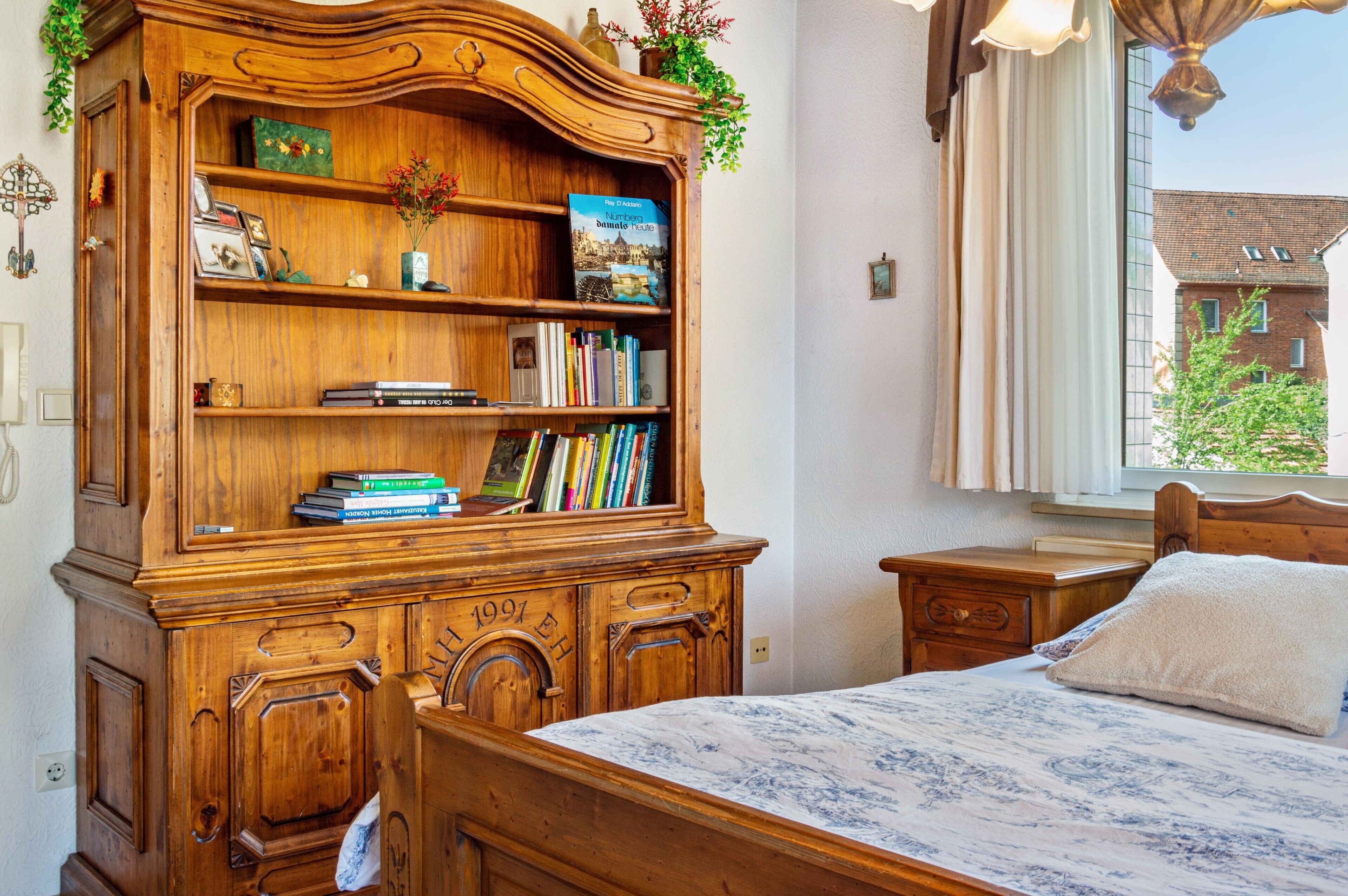 A bedroom with a wooden bookcase, a bed, a nightstand, and a window with a view of buildings.