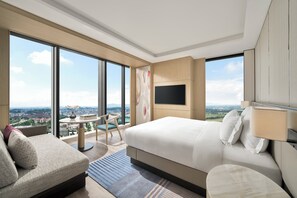 Habitación de hotel moderna con una cama grande, un televisor en la pared, una mesita pequeña y vistas de la ciudad a través de amplias ventanas.