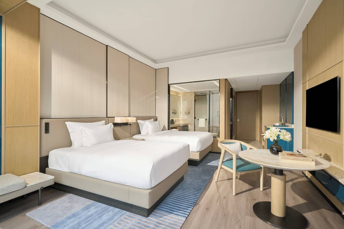 Ett modernt hotellrum med två sängar, ett litet bord, en TV och ett badrum som syns genom en glasdörr.
