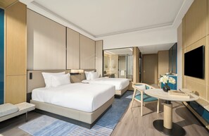 Habitación de hotel moderna con dos camas, una mesita, un televisor y un baño visible a través de una puerta de vidrio.
