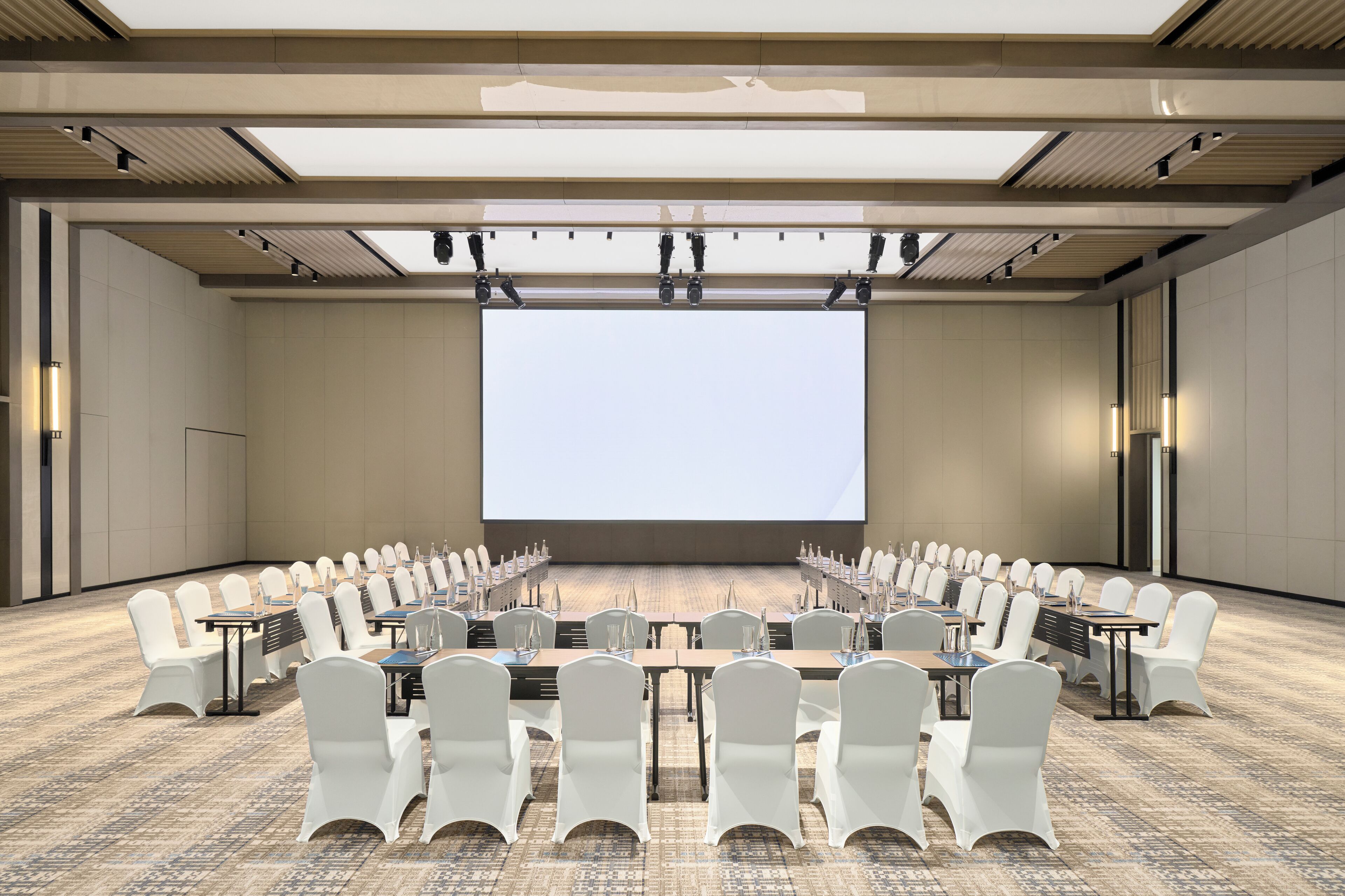 Un salón de conferencias con una pantalla grande, filas de sillas blancas y mesas con botellas de agua.