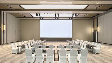 A conference room with a large screen, rows of white chairs, and tables with water bottles.