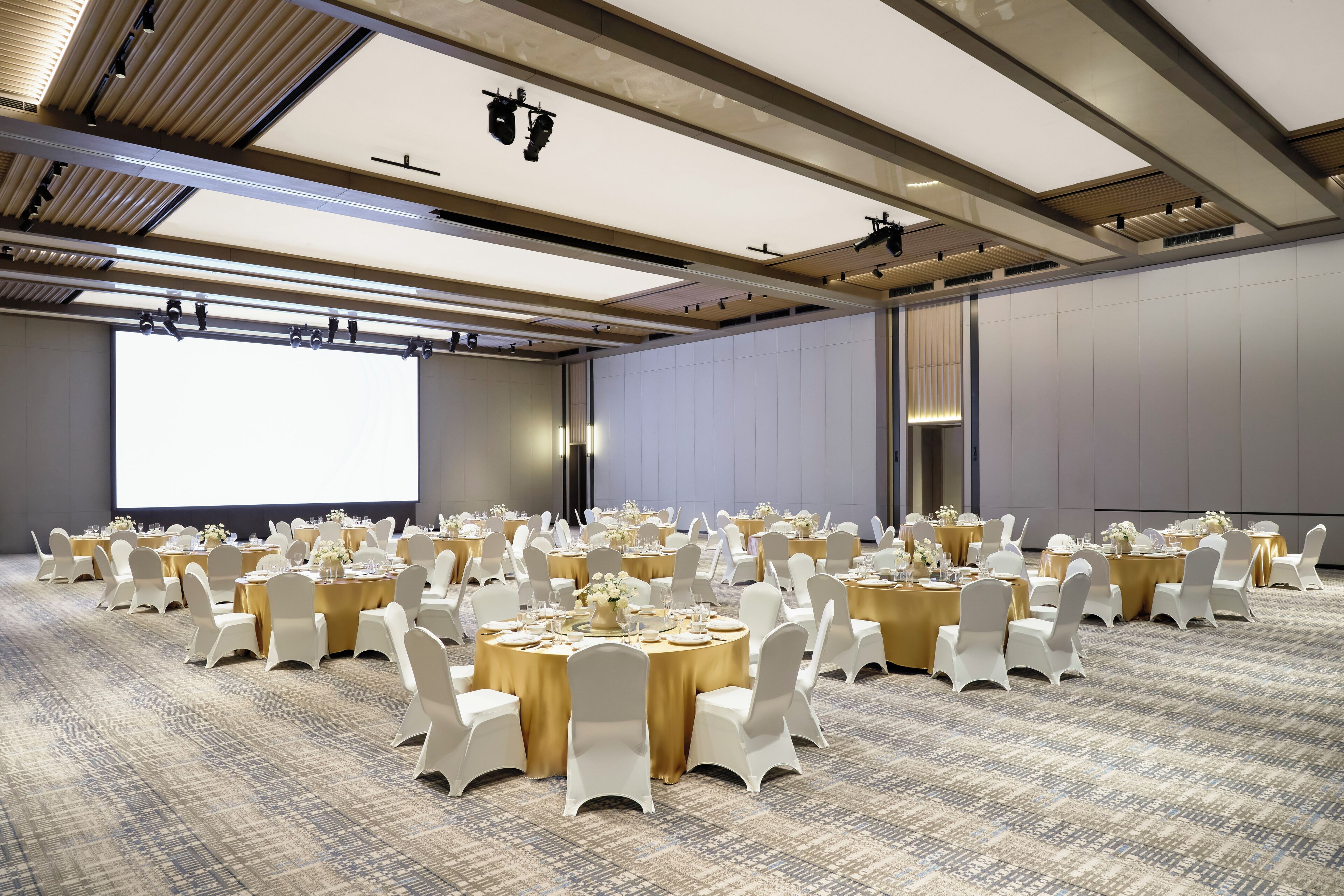 A large conference room with multiple round tables, white chairs, and a large screen at the front.