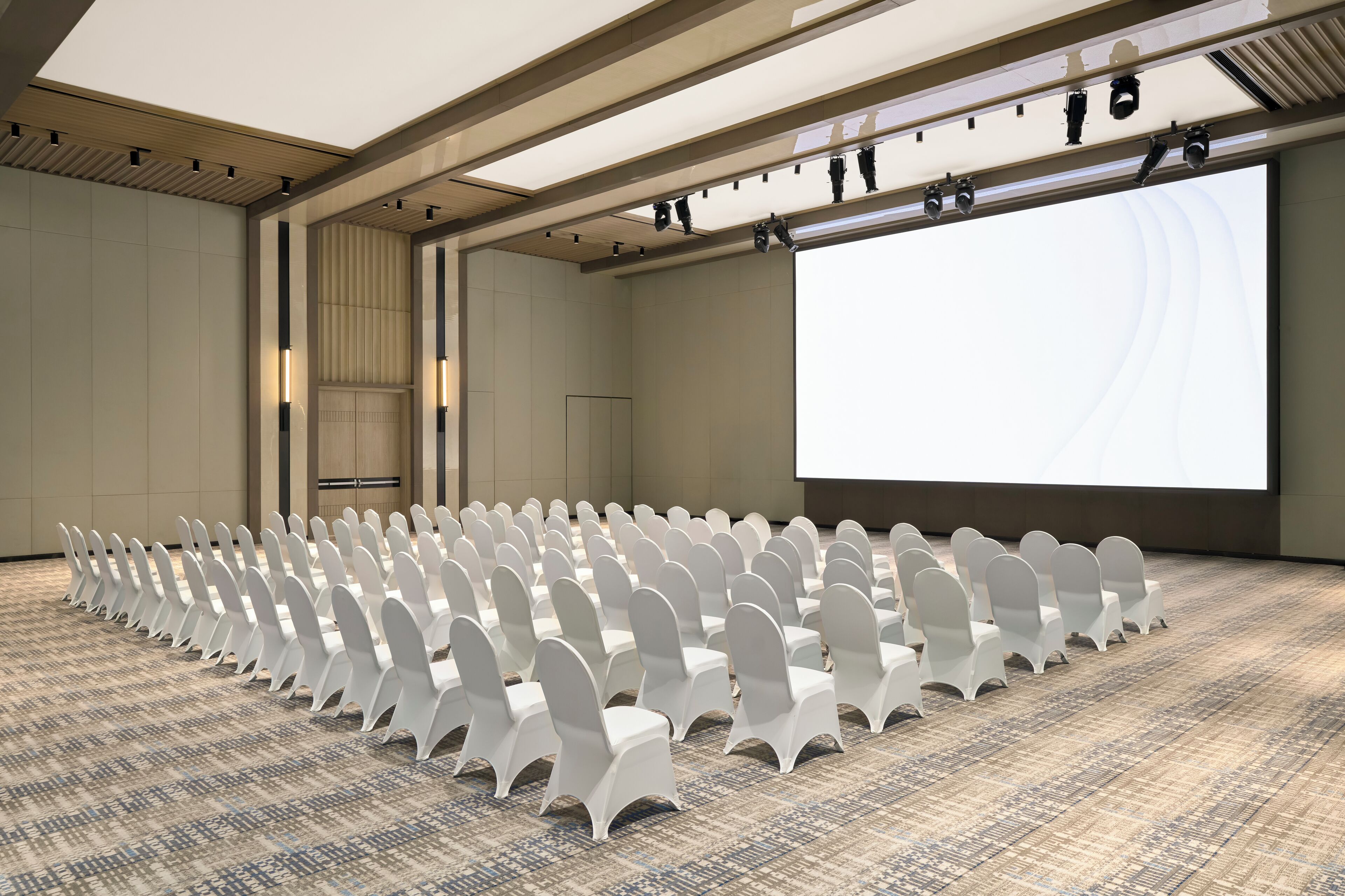 Une salle de conférence avec des rangées de chaises blanches, un grand écran et une moquette à motifs.