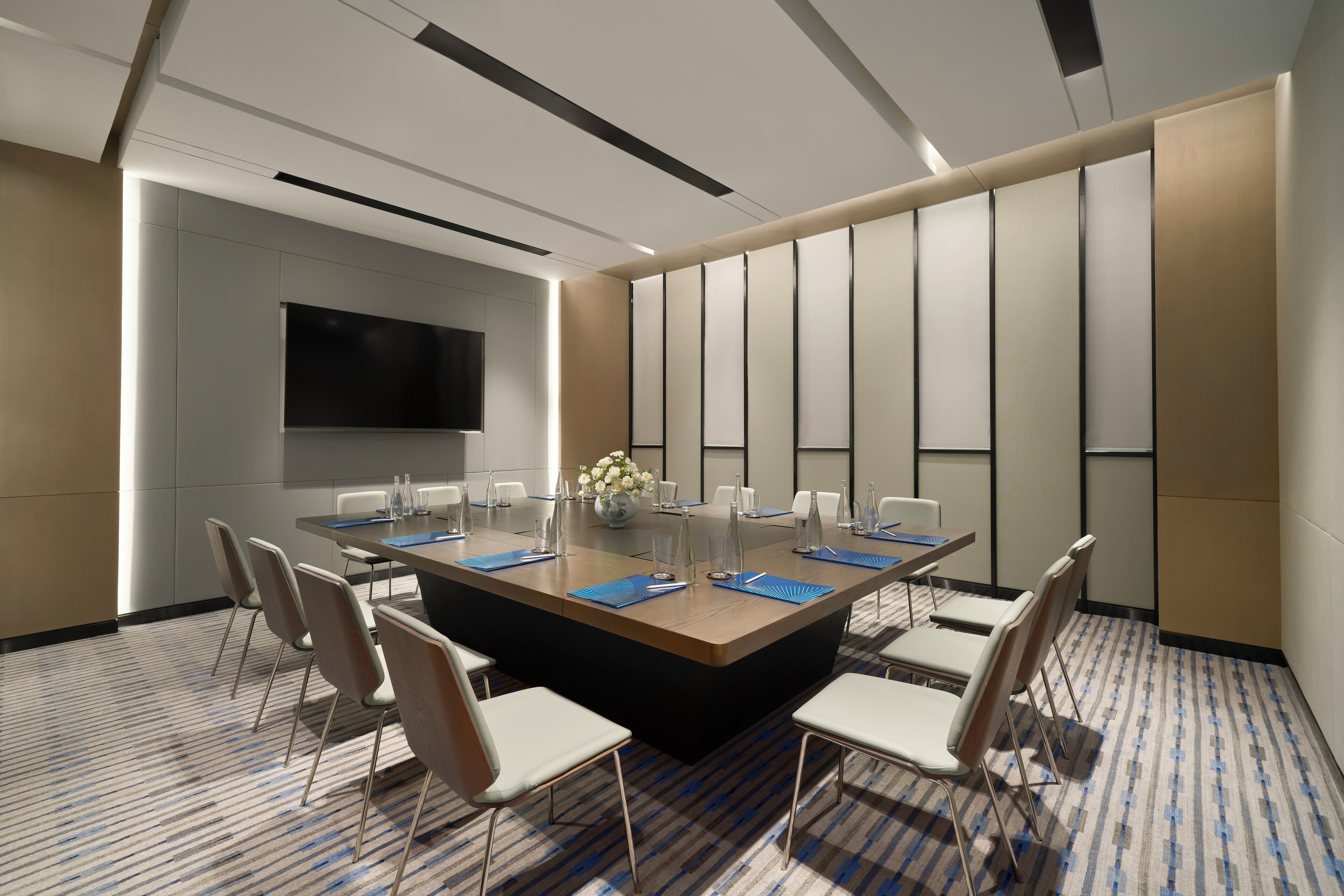 A conference room with a large rectangular table, white chairs, a flat-screen TV, and a patterned carpet.